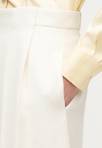 Χέρι στην τσέπη λευκού παντελονιού ψηλής μέσης με φαρδιά μέση, σε συνδυασμό με ανοιχτόχρωμο κίτρινο πουκάμισο με μακριά μανίκια και κουμπωτά μανσέτα.