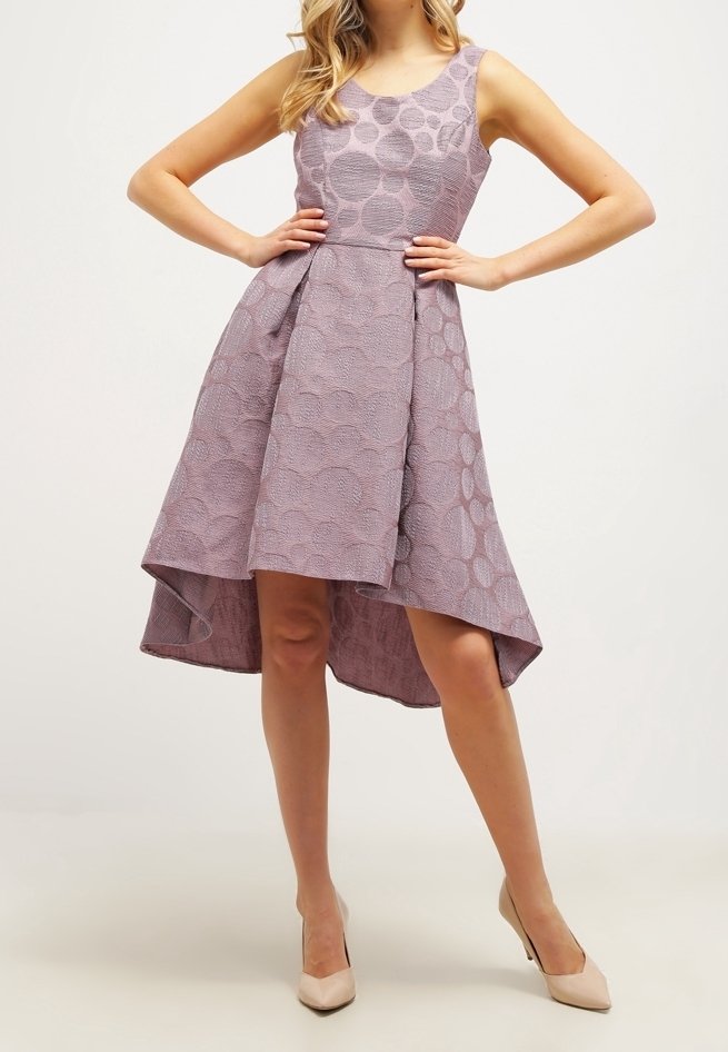 Robe sans manches lilas avec des motifs circulaires texturés, corsage ajusté et ourlet asymétrique évasé. Associée à des chaussures à talons nude.