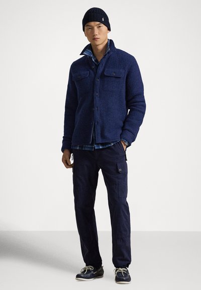 Marineblaues Hemdjacke aus Wollmischung mit zwei aufgesetzten Brusttaschen mit Knöpfen, darüber ein kariertes Hemd, kombiniert mit marineblauen Cargo-Hosen und dunklen Schuhen.