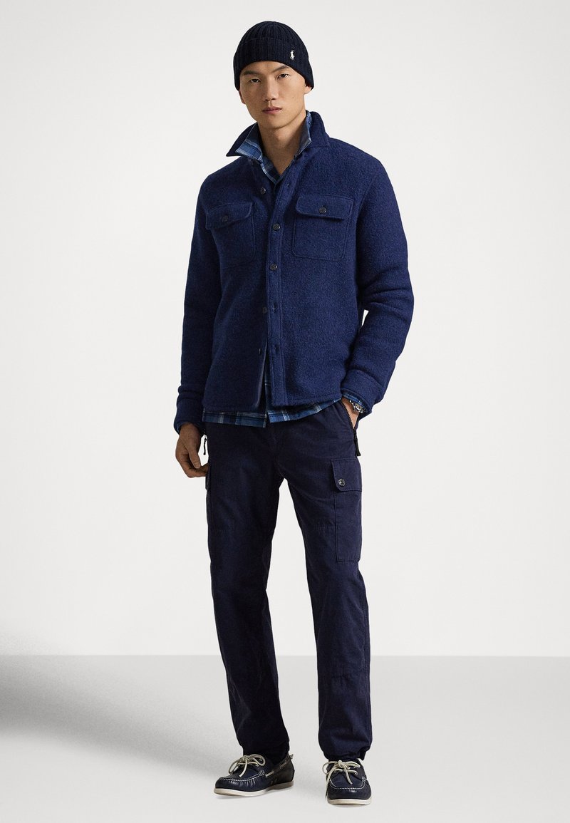 Veste-chemise bleu marine en mélange de laine avec deux poches poitrine à boutons, portée sur une chemise à carreaux, associée à un pantalon cargo bleu marine et des chaussures foncées.
