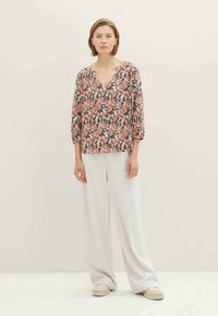 Donna in piedi con una blusa floreale a scollo a V, pantaloni bianchi a gamba larga e scarpe beige, di fronte a uno sfondo chiaro e semplice.