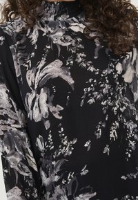 Schwarze Bluse mit Floralprint in Grautönen und Weiß. Mit einem hohen, gerüschten Kragen und strukturiertem, durchscheinendem Stoff.