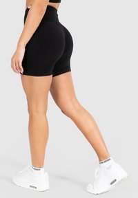 Czarne szorty z wysokim stanem charakteryzują się gładką teksturą i dopasowanym krojem, zestawione z białymi butami sportowymi i kontrastującymi czarnymi akcentami.