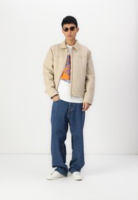 Chaqueta beige con cuello, cierre frontal y detalles de costura, combinada con una camiseta gráfica blanca, jeans de mezclilla sueltos y zapatillas blancas.