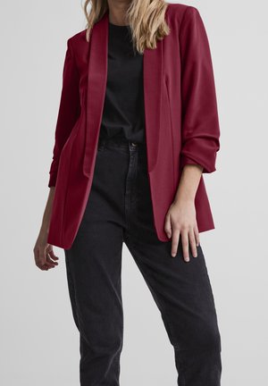 Blazer rouge avec un design structuré, doté d'un col châle et de poches avant, associé à un t-shirt noir et à un jean foncé. Texture lisse.