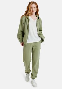 Olivengrüner Baumwollhoodie mit Fronttaschen, kombiniert mit passenden Sweatpants und weißen Sneakers, bietet einen lockeren Schnitt und eine weiche Textur.