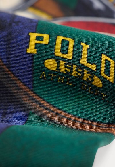 Близък план на плат с жълт текст "Polo 1993 ATHL. DEPT." на зелен и син фон, показващ детайлна плетена текстура.