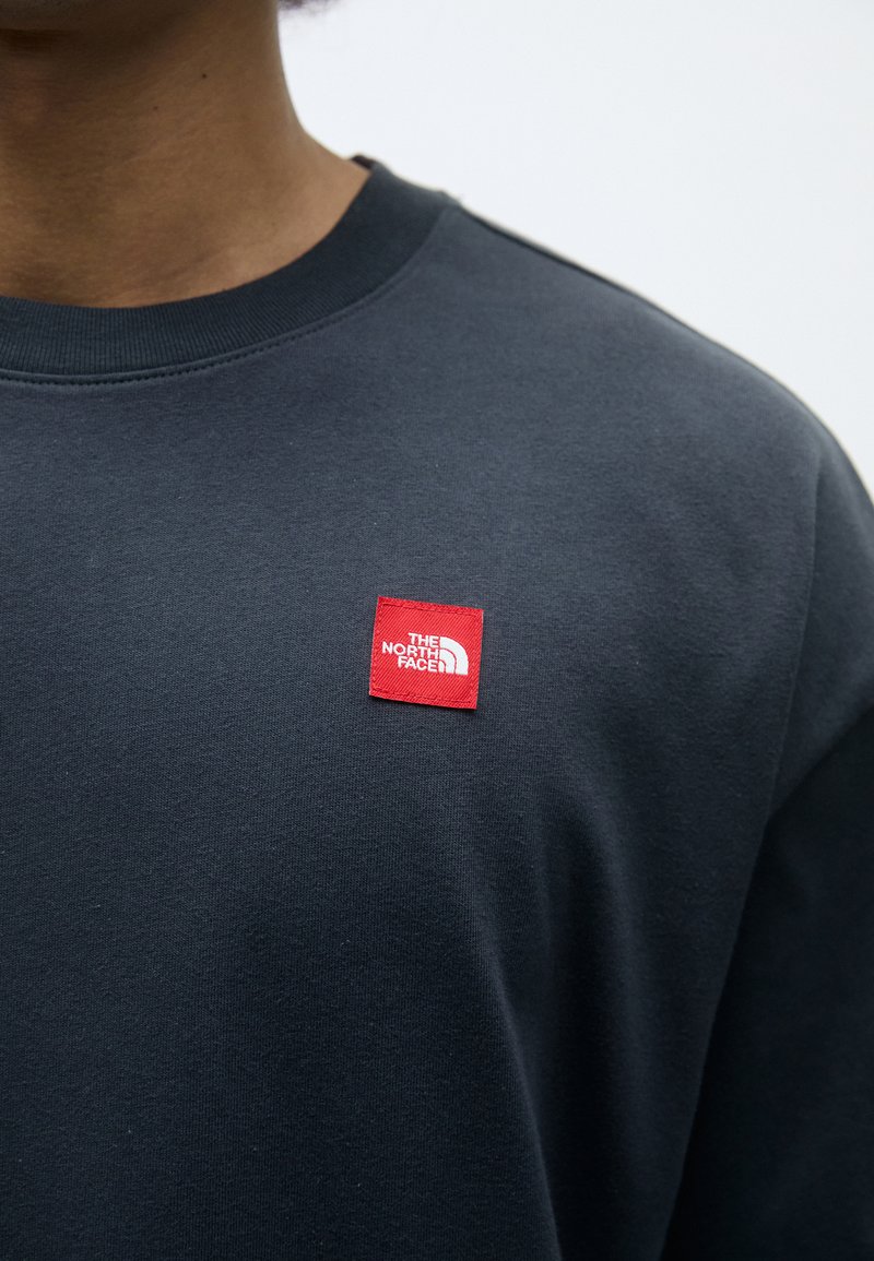 Felpa collo crew navy con un patch quadrato rosso etichettato "THE NORTH FACE" sull'area del petto sinistro. Texture in cotone liscia.