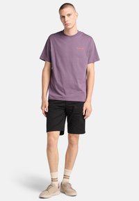 Camiseta de algodón morada con logo naranja pequeño, acompañada de pantalones cortos negros. El modelo lleva zapatos beige y calcetines crema con rayas marrones.