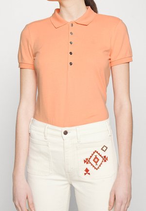 Frau trägt ein kurzärmeliges pfirsichfarbenes Poloshirt und hochtaillierte weiße Hose mit roter geometrischer Stickerei auf der rechten Tasche.