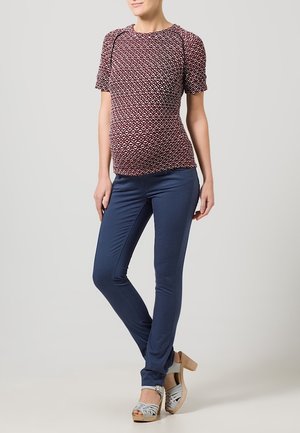 Blusa de maternidad con patrón geométrico multicolor, mangas cortas; combinada con pantalones ajustados en azul marino y sandalias de cuña grises.