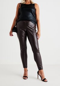 Sort ærmeløs top med en blank finish, parret med tætsiddende brune faux læderbukser og sorte strophæle.