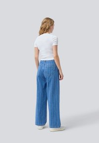 Weite blaue Denim-Jeans mit vertikalen weißen Streifen, ausgestattet mit zwei Gesäßtaschen. Tragen Sie dazu ein eng anliegendes weißes T-Shirt mit kurzen Ärmeln und weiße Schuhe.