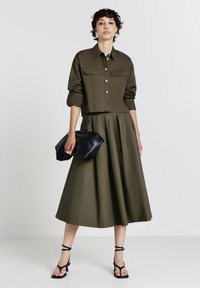 Camicetta verde oliva corta con due tasche frontali, abbinata a una gonna lunga dello stesso colore, che tiene una clutch nera e indossa decolletè nere con cinturino.