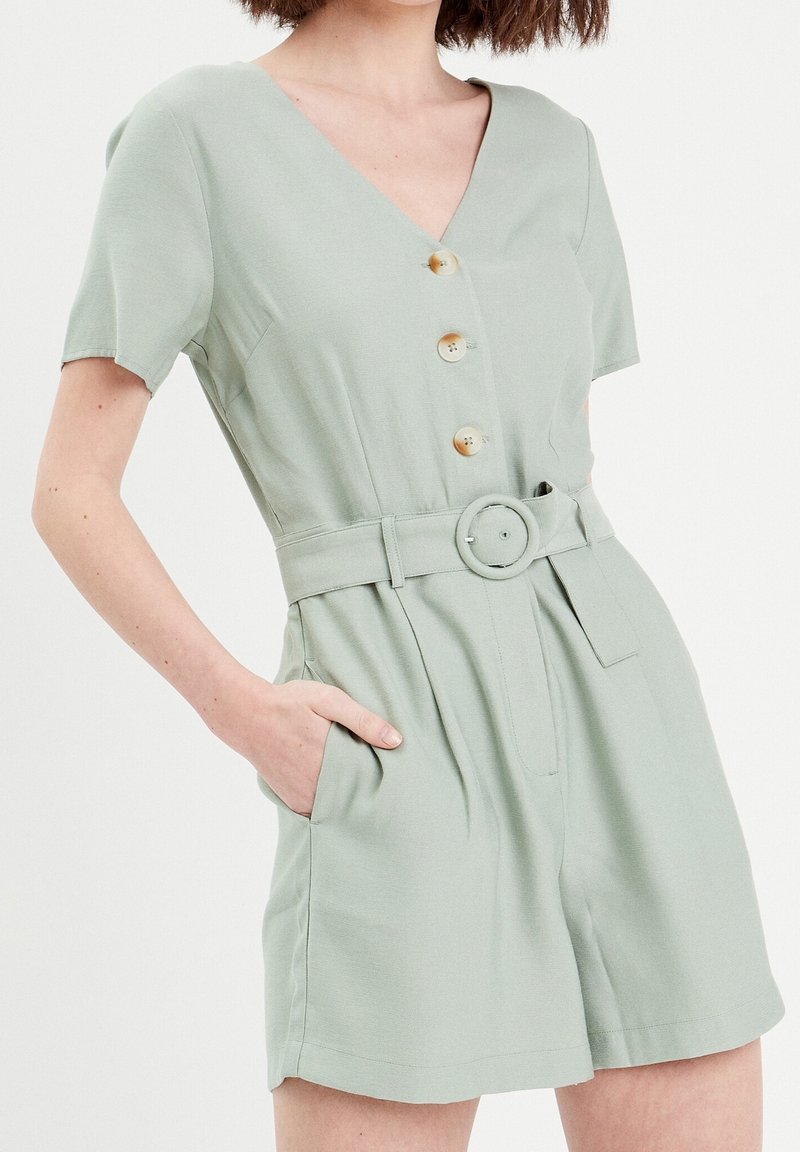 Femme portant une combinaison courte vert clair à manches courtes avec ceinture, boutons et poches, main reposant dans la poche droite, visible du cou à la mi-cuisse.