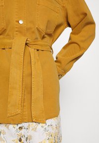 Hořčicově žlutá džínsová bunda s opaskem v pase, s knoflíkovými klopovými kapsami a uvolněným střihem. Obsahuje viditelné švy a měkkou texturu.