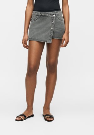 Femme portant un haut noir sans manches, une mini-jupe grise asymétrique boutonnée et des sandales plates noires avec détail chaîne dorée.