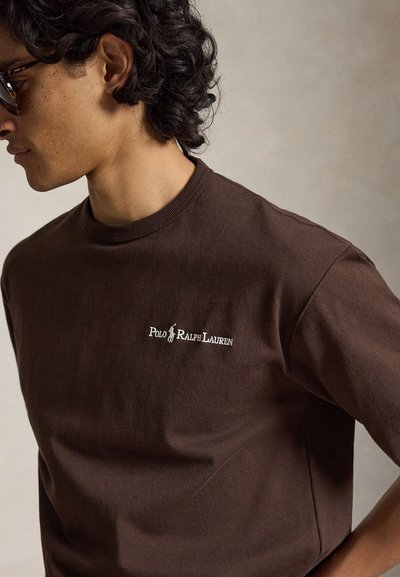 Άνδρας που φοράει καφέ μπλούζα Polo Ralph Lauren και γυαλιά ηλίου με σχέδιο χελώνας, γυρισμένος πλαγίως προς απλό φόντο.