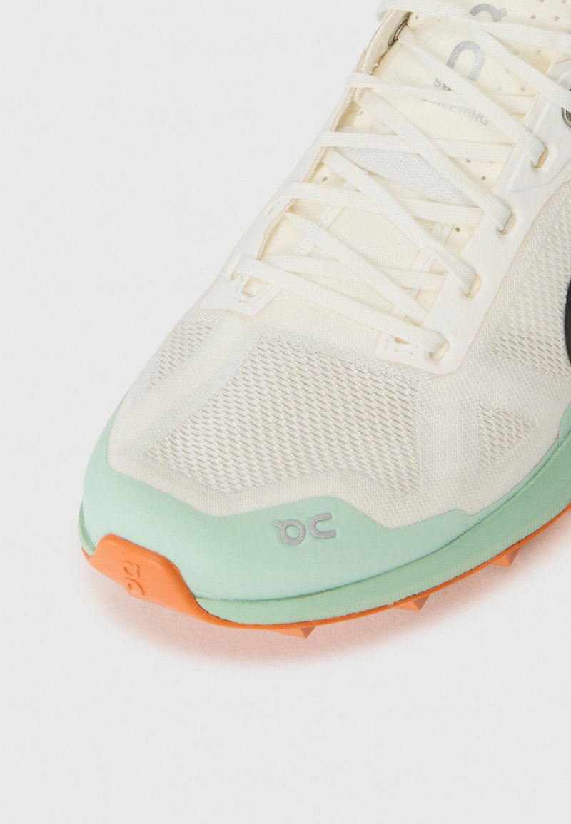 Zapatilla deportiva ligera en blanco y verde menta, con un upper de malla texturizada, acento naranja en la suela exterior y detalle de logo plateado.