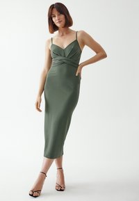 Olivgrünes Midi-Kleid mit figurbetontem Schnitt, V-Ausschnitt und Spaghettiträgern. Besitzt ein gedrehtes Detail an der Taille. Glatter, dehnbarer Stoff.