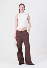 Bílý bezrukávový top s detailem mašle na boku, kombinovaný s hnědými kalhotami širokého střihu. Modelka drží malou bílou kabelku; špičaté černé boty.