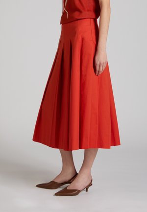 Frau trägt einen plissierten, leuchtend orangefarbenen Midirock, kombiniert mit braunen, spitz zulaufenden Slingback-Schuhen mit niedrigem Absatz vor einem einfarbigen Hintergrund.