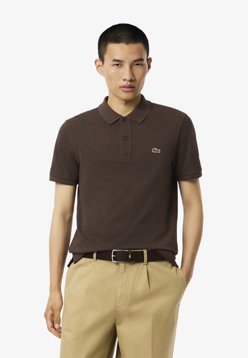 Brun kortärmad polo tröja med krage, med en liten grön Lacoste-logotyp. Bärs med beige byxor och ett brunt bälte.