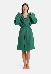 Accappatoio verde in spugna con maniche lunghe, vita attillata e due tasche frontali; presenta una finitura testurizzata e una chiusura con legaccio.