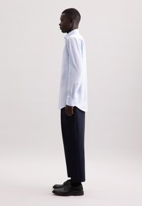 Uomo di profilo vestito con una camicia a righe blu chiaro e bottoni, pantaloni scuri e scarpe eleganti nere, davanti a uno sfondo uniforme.