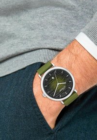 Runde Silberuhr mit schwarzem, strukturiertem Zifferblatt, grünen Akzenten und einem grünen Stoffarmband. Am Handgelenk getragen, unter einem grauen Pullover versteckt.