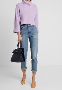 Liliowa sweter z golfem o prążkowanej fakturze, dżinsy w średnim odcieniu z mankietami, granatowa torebka z detalem w kształcie kokardy i beżowe szpilki w szpic.