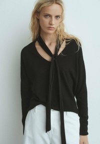 Maglione nero lavorato a maglia con scollo a V e dettaglio con fiocco, con maniche lunghe e vestibilità rilassata, abbinato a pantaloni bianchi.