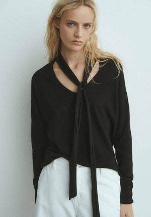 Pull en tricot noir avec un col en V et un détail à nouer, doté de manches longues et d'une coupe décontractée, associé à un pantalon blanc.