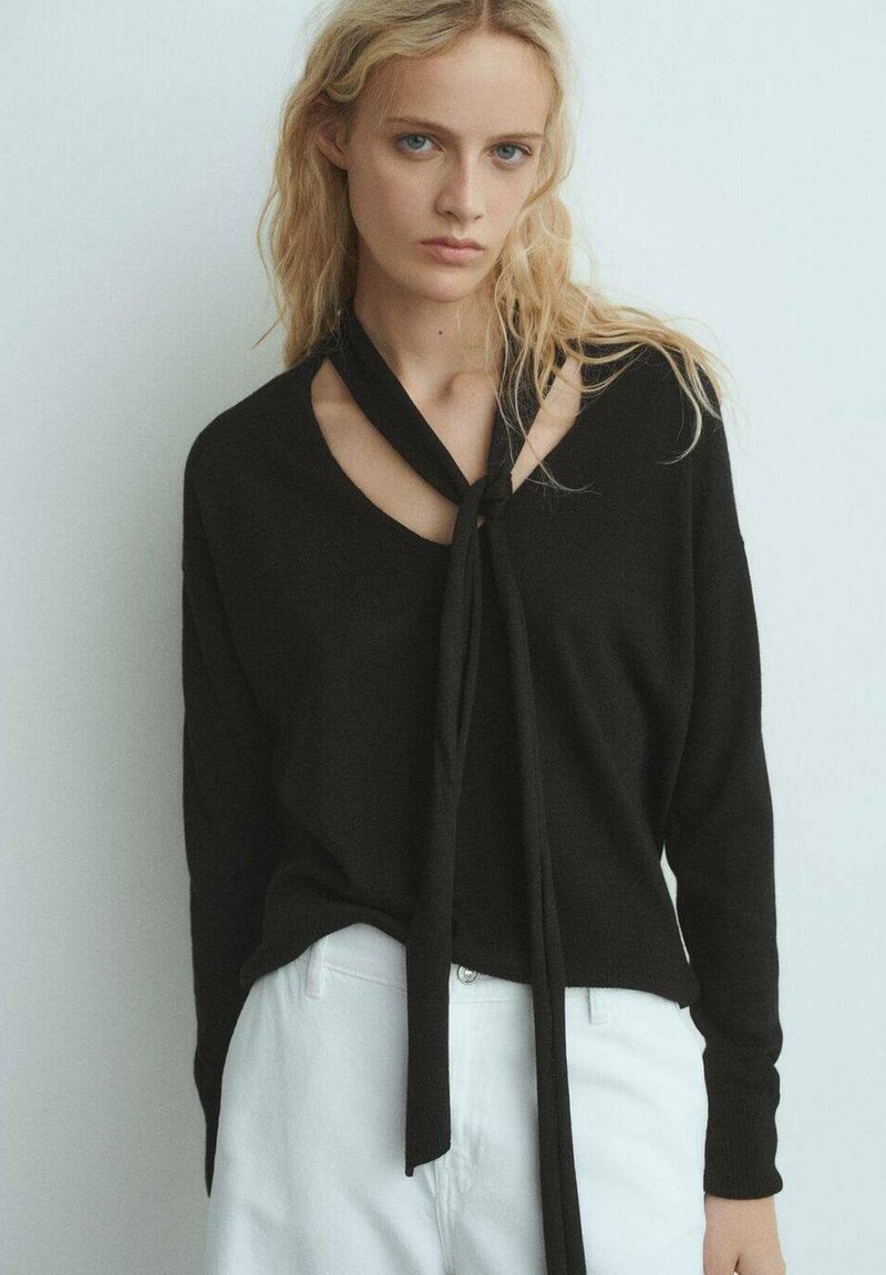 Maglione nero lavorato a maglia con scollo a V e dettaglio con fiocco, con maniche lunghe e vestibilità rilassata, abbinato a pantaloni bianchi.