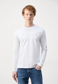 Hvid langærmet t-shirt i blød stof med rund halsudskæring og et lille logo på brystet, iført med blå denim jeans.