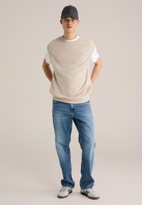 Šviesiai rudas megztas berankovis megztinis dėvimas ant baltos marškinėlių, derinamas su šviesiai mėlynomis tiesių kojų džinsais ir pilkais kedais su užlenktu viršumi. Neutralus fonas.