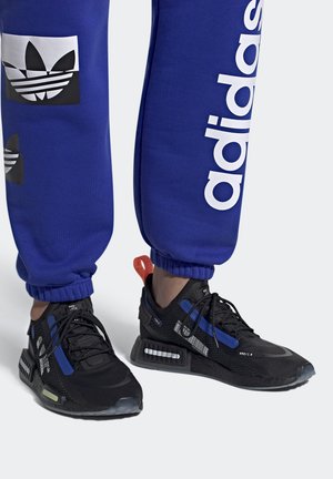 Henkilö, joka pukeutunut sinisiin Adidas-housuihin ja mustiin Adidas-tossuihin, joissa on sinisiä ja oransseja yksityiskohtia, seisoo valkoisella pinnalla.