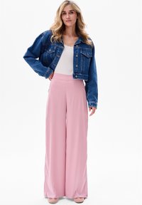 Jeansowa kurtka z guzikami, biała prążkowana bluzka i jasnoróżowe szerokie spodnie. Gładki materiał; modelka stoi na tle białym.