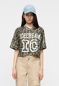 Νεαρή γυναίκα που φοράει μπλε καπέλο με την επιγραφή "HOT", κοντό τοπ με σχέδιο λεοπάρδαλης και γράμματα "ICECREAM IC" και μπεζ παντελόνι, μπροστά από απλό φόντο.