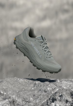 Scarpa da trail Merrell grigia con suola spessa sospesa sopra un terreno roccioso, piccole particelle di terra cadono intorno, sfondo sfocato isolato.