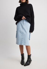 Una donna che indossa un maglione nero a collo alto, una gonna midi in denim blu chiaro con tasche laterali e stivali neri a punta fino al polpaccio, in piedi su uno sfondo neutro.