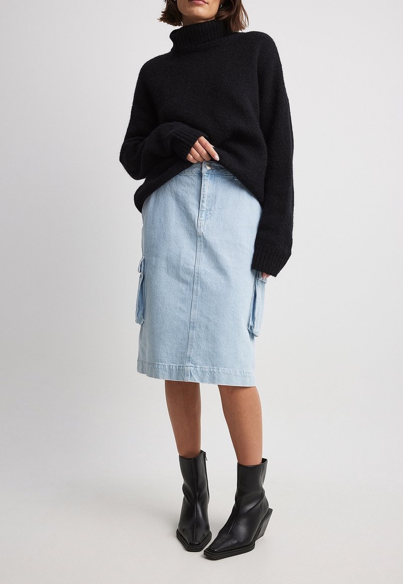 Una donna che indossa un maglione nero a collo alto, una gonna midi in denim blu chiaro con tasche laterali e stivali neri a punta fino al polpaccio, in piedi su uno sfondo neutro.