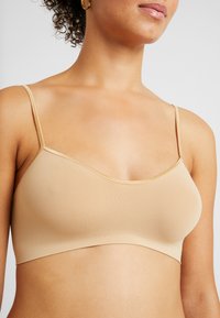 Bralette leggero nudo con spalline sottili e regolabili, una texture liscia e una sottile rifinitura metallica lungo il scollo e la fascia.