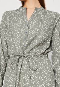 Femme portant une robe verte à boutons avec des motifs floraux blancs, à manches longues et une ceinture nouée à la taille.