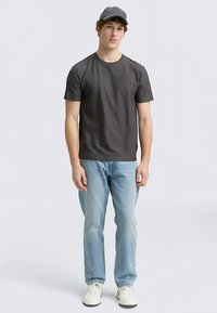 Tânăr cu un tricou gri închis, blugi bleu deschis, adidași albi și șapcă gri de baseball, stând pe un fundal simplu deschis la culoare.