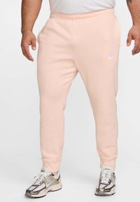 Pantalones de chándal rosa claro con cinturilla elástica, piernas ajustadas y un pequeño logo blanco en la cadera izquierda; combinados con zapatillas deportivas.