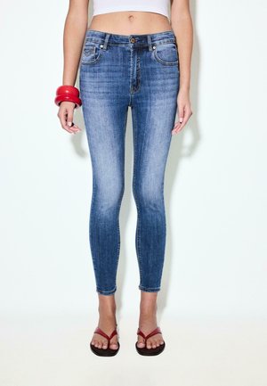 Kvinde iført blå skinny jeans med mellemhøj talje, hvid crop top, røde armbånd på venstre håndled og røde flip-flops på hvid baggrund.