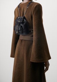 Sac à dos en cuir noir avec texture matelassée, accents en chaîne et poche avant, porté avec une robe à manches longues, marron clair et duveteuse.