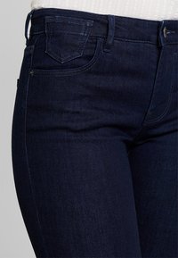 Primer plano de unos vaqueros de denim azul oscuro llevados con una camiseta blanca de canalé metida por dentro, mostrando los detalles de los bolsillos frontales y laterales, así como la cinturilla.