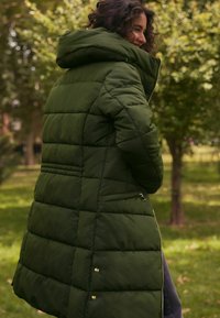 Zeleni kaput s podstavom i kapuljačom, s prešanim uzorkom, bočnim džepovima sa zatvaračima i zlatnim detaljima. Mekana, teksturirana tkanina.